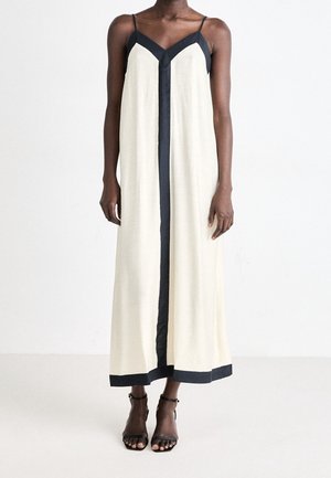 Femme portant une robe longue crème sans manches avec des bordures noires le long de l'encolure, du centre et de l'ourlet, ainsi que des talons noirs à lanières.