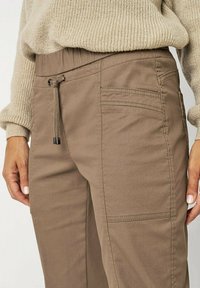 Braune Baumwollmischungshosen mit elastischem Bund, verstellbarem Kordelzug und seitlichen Taschen. Texturierte Oberfläche und maßgeschneiderte Fit-Details sind sichtbar.