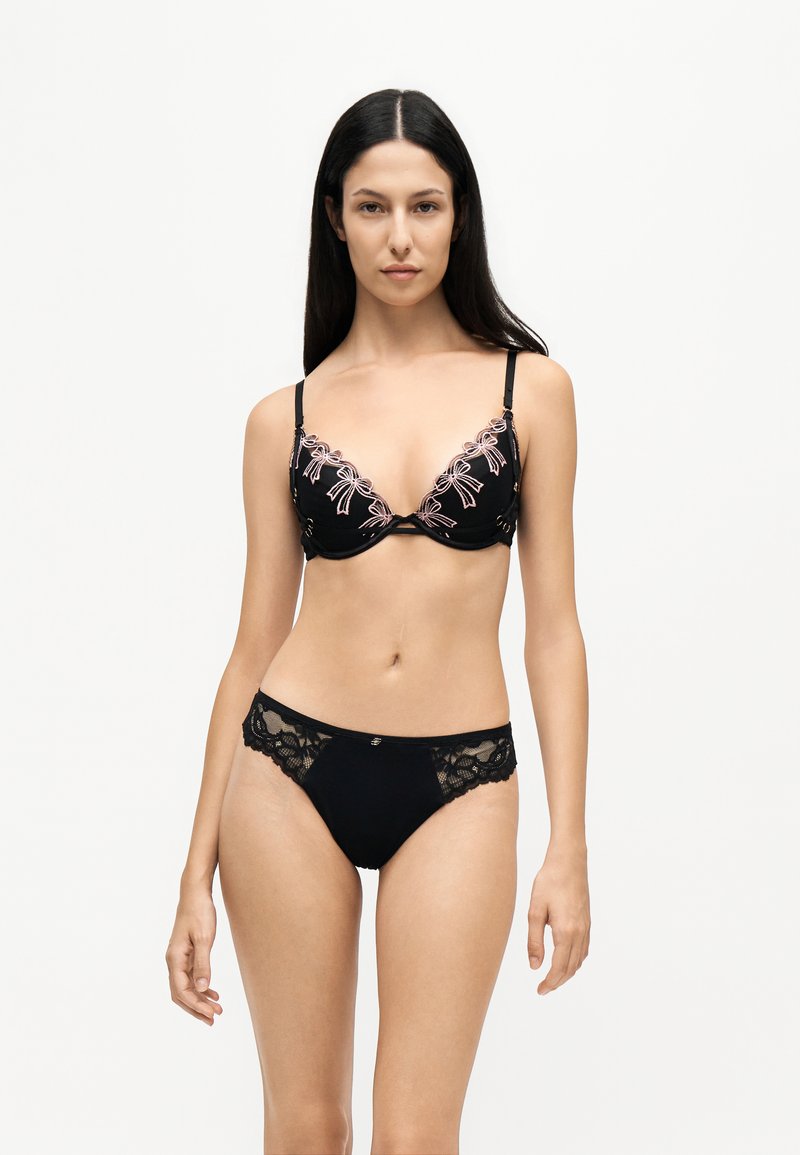 Femme aux longs cheveux foncés portant un soutien-gorge noir avec broderie rose et une culotte noire en dentelle assortie, debout devant un fond uni.