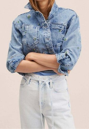 Kobieta ze skrzyżowanymi ramionami, ubrana w jasnoniebieską haftowaną kurtkę dżinsową i jasnoniebieskie jeansy z wysokim stanem z przewiązanym paskiem.