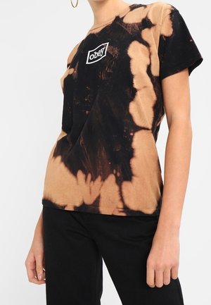 Person trägt ein schwarz-beiges Batik-T-Shirt mit "obey"-Logo auf der Brust und schwarze Hose, steht vor neutralem Hintergrund.