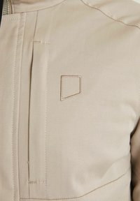 Beige jacka med vertikal dragkedja ficka framtill och en liten broderad geometrisk logotyp på bröstet.