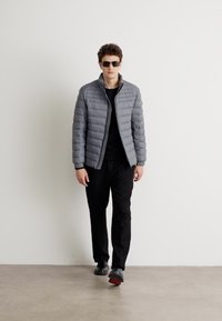 Gray quilted puffer jacket with a stand collar, paired with a black top and trousers. The model wears dark sunglasses and black shoes.