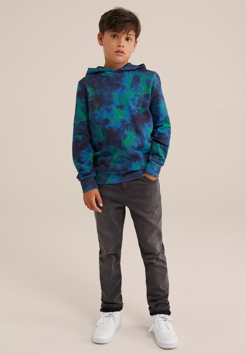 Sudadera con capucha en un estampado abstracto azul y verde, con cordones en la capucha. Combinada con jeans gris oscuro y zapatillas blancas.