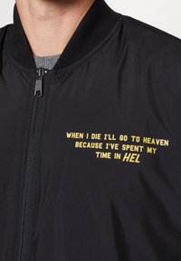 Giacca bomber nera con zip frontale. Il testo giallo recita: "QUANDO MORIRÒ ANDRÒ IN CIELO PERCHÉ HO TRASCORSO IL MIO TEMPO IN INFERNO."