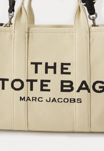 Beige canvas toteväska med svart text "THE TOTE BAG" och "MARC JACOBS." Har dubbla handtag och dragkedjestängning, strukturerad yta.