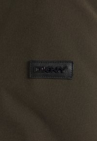 La tela de oliva oscura con una textura suave presenta un parche de cuero negro con el logo de DKNY cosido, mostrando letras en relieve y una forma rectangular.