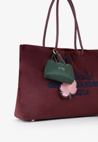 Borsa a tracolla in velluto bordeaux con testo blu ricamato, dotata di una piccola pochette in pelle verde e di un charm floreale rosa attaccato.