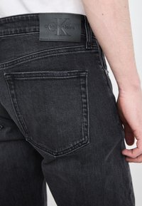 Fekete farmernadrág látható hátsó zsebbel és texturált bőr derékkal, amelyen a "Calvin Klein Jeans" felirat van belepréselve. Testhezálló dizájn.