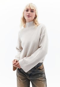 Hellbeige, gerippter Rollkragenpullover mit weiten Ärmeln, kombiniert mit ausgewaschenen blauen Jeans. Die Textur ist weich und hat eine lässige Passform.