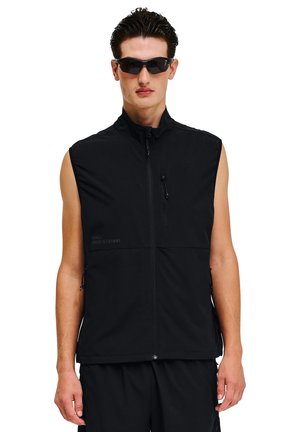 Mężczyzna ubrany w czarną bezrękawnik na zamek błyskawiczny i czarne okulary przeciwsłoneczne, stoi na białym tle.