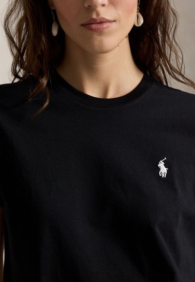 Γυναίκα που φοράει μαύρο μπλουζάκι με στρογγυλή λαιμόκοψη και μικρό λευκό λογότυπο παίκτη πόλο στο στήθος, με σκουλαρίκια από κοχύλια που κρέμονται, εμφανιζόμενη από τον λαιμό μέχρι το στήθος.