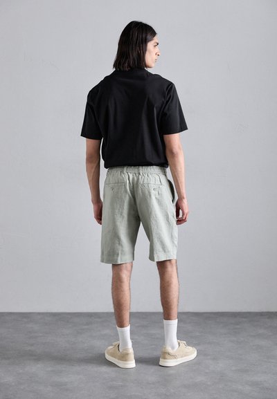 Schwarzes kurzärmeliges Shirt kombiniert mit hellgrünen Shorts mit Kordelzug, zwei Gesäßtaschen und lockerer Passform. Sneakers und Socken inklusive.
