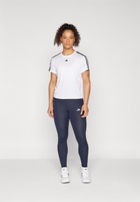 T-shirt bianco a maniche corte con strisce nere sulle spalle, abbinato a leggings blu navy e scarpe da ginnastica bianche, con loghi del marchio.