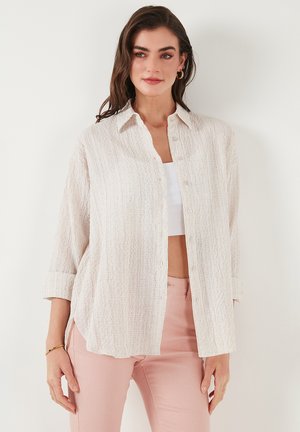 Light beige button-up shirt with a textured fabric, long sleeves rolled at cuffs, and a relaxed fit, paired with pastel pink pants.