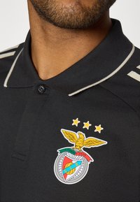Czarna koszulka polo z dwukolorowym kołnierzykiem i wyraźnym haftowanym logo klubu przedstawiającym orła oraz trzy gwiazdy nad nim.