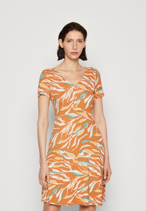 Γυναίκα με σκούρα μαλλιά, φορεμένη με ένα κοντομάνικο πορτοκαλί φόρεμα με μπλε, λευκά και κίτρινα αφηρημένα σχέδια φύλλων, σε απλό φόντο.