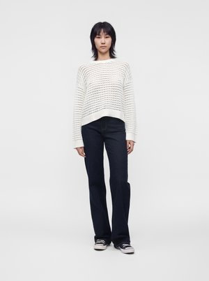 Personne debout de face, portant un pull en maille blanc ample, un jean foncé et des baskets noires et blanches sur un fond uni clair.