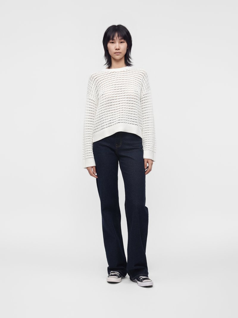 Personne debout de face, portant un pull en maille blanc ample, un jean foncé et des baskets noires et blanches sur un fond uni clair.