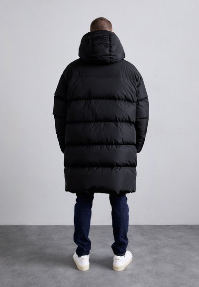 Une personne se tient face à un mur, portant un long manteau rembourré noir, un jean sombre et des baskets blanches avec du texte sur les talons.