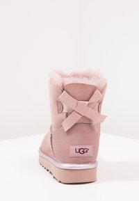 Růžová semišová kotníková bota s měkkou kožešinovou podšívkou, ozdobnou mašlí na zadní straně a strukturovanou gumovou podrážkou. Logo UGG na patě.