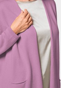 Hellblauer Cardigan mit weicher Textur, offenem Vorderteil und zwei Taschen, kombiniert mit einem glatten, silbernen Oberteil. Eine Hand hält den Cardigan sanft.
