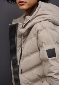 Beige puffer bunda s kapucí, elastickými šňůrkami a černou nášivkou na rukávu. Texturovaný prošívaný design s lesklým povrchem.
