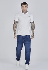 Valge ümarakaelusega t-särk, millel on väike must logo, kombineeritud siniste teksapükstega, mis on lõdva lõike ja peene tekstuuriga. Valged vabaajajalatsid.