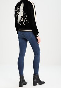 Giubbotto bomber nero con ricami di gru e design floreali, strisce laterali crema, polsini a coste, indossato su jeans blu e stivaletti neri.