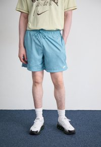 Bermudas atléticas de cor azul clara com uma cintura elástica, logótipo da Nike em branco. Combinadas com uma t-shirt amarela pálida e ténis brancos com detalhes verdes.