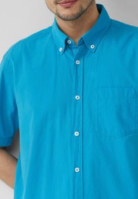 Kurzärmliges türkises Button-up-Hemd aus strukturiertem Stoff, mit weißen Knöpfen und einer aufgesetzten Tasche auf der linken Seite.