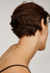 Pendientes de aro dorados con un diseño texturizado, combinados con un collar de cadena delgada y una delicada sarta de perlas, sobre piel desnuda con pelo corto castaño.