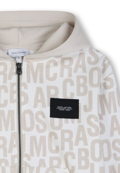 Sweat à capuche zippé beige et blanc avec un motif de texte "MARC JACOBS" répété et un patch noir portant l'inscription "MARC JACOBS LOCATION : N.Y.C. YEAR : 1984".