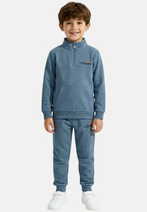 Jeune garçon souriant portant un sweat-shirt à fermeture éclair bleu et un pantalon de survêtement assorti avec des baskets blanches, debout devant un fond uni.