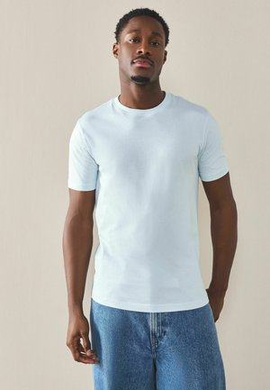 Bărbat cu păr scurt și creț, purtând un tricou albastru deschis și blugi albaștri, stând în picioare pe un fundal simplu bej.