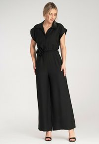 Czarny kombinezon z krótkimi, podwiniętymi rękawami, zapięciem na guziki z przodu i paskiem z materiałowym kwiatem. Posiada szerokie nogawki i gładką, lekką strukturę.
