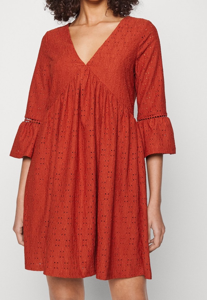Femme portant une robe ample orange rouille en tissu ajouré, encolure en V et manches évasées trois-quarts avec détails en dentelle.