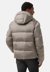 Grå jakke med dun, der har hætte, med en kviltet design, glat tekstur og logo på ryggen. Båret over mørke jeans.