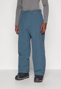 Muškarac u širokim plavim hlačama sa poja�čanim šavovima, crnim cipelama i sivoj majici dugih rukava, stoji prema jednostavnoj pozadini.