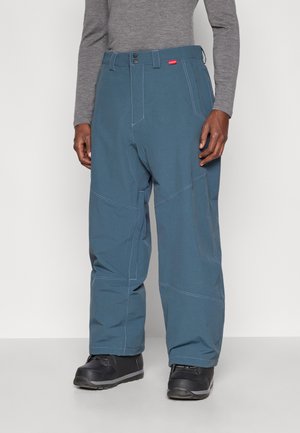 Egy férfi laza kék nadrágot visel, megerősített varrásokkal, fekete cipőkkel és szürke, hosszú ujjú felsővel, egy sima háttér előtt áll.