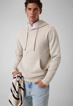 Junger Mann trägt einen beigen Hoodie über einem weißen Hemd, hellblaue Jeans, hält einen cremefarbenen Schal mit schwarzen Streifen in einer Hand.