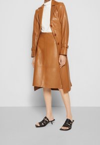 Trench-coat marron en cuir avec une taille ajustée, assorti à une jupe midi en cuir assortie. Sandales noires à bout ouvert avec trois brides.