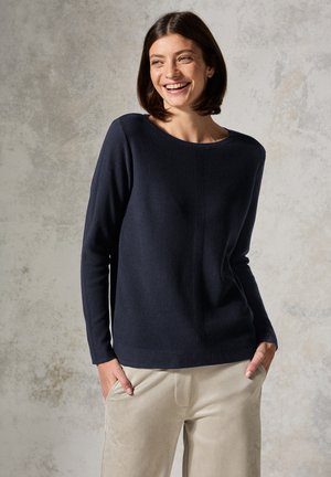 Pull ribbed marine avec un col rond et des manches longues, présentant un détail de couture vertical. Associé à un pantalon beige à texture douce.