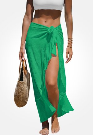 Gonna verde avvolgente con chiusura a cravatta e spacco laterale, realizzata in tessuto leggero. Abbinata a una borsa intrecciata e indossata a piedi nudi.