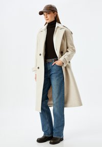 Abrigo de mezcla de lana beige con cuello de muesca, usado sobre un suéter de cuello alto oscuro y jeans rectos azules. Incluye una gorra de cuero marrón y botas oscuras.