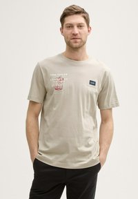 Camiseta de algodón beige claro con cuello redondo, mangas cortas y un diseño de texto impreso en la parte frontal. Corte holgado, textura suave.