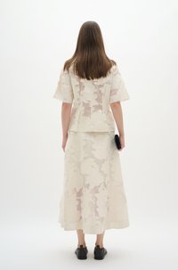 Helles, leichtes, weißes Kleid mit floralem Design; hat kurze Ärmel und eine fließende Silhouette. Schwarze Schuhe und eine Clutch unterstreichen den Look.
