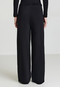 Schwarze weite Hosen mit elastischem Bund und glatter Textur, im vollen Längenformat, von hinten gezeigt.