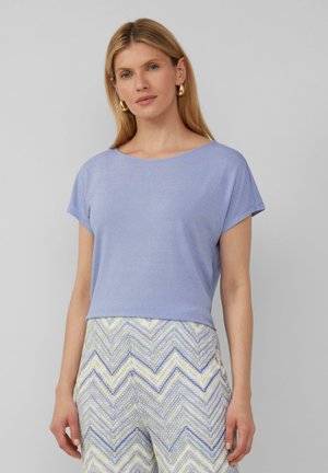 Haut mauve clair à manches courtes avec un col rond, fabriqué dans un tissu lisse et légèrement brillant. Associé à un pantalon multicolore à motifs.