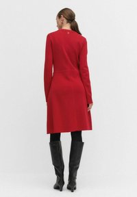 Rode langemouwen jurk met een aansluitend lijfje en een uitlopende rok, voorzien van een gladde textuur en een knoopdetail op de rug, gecombineerd met zwarte laarzen tot de knie.